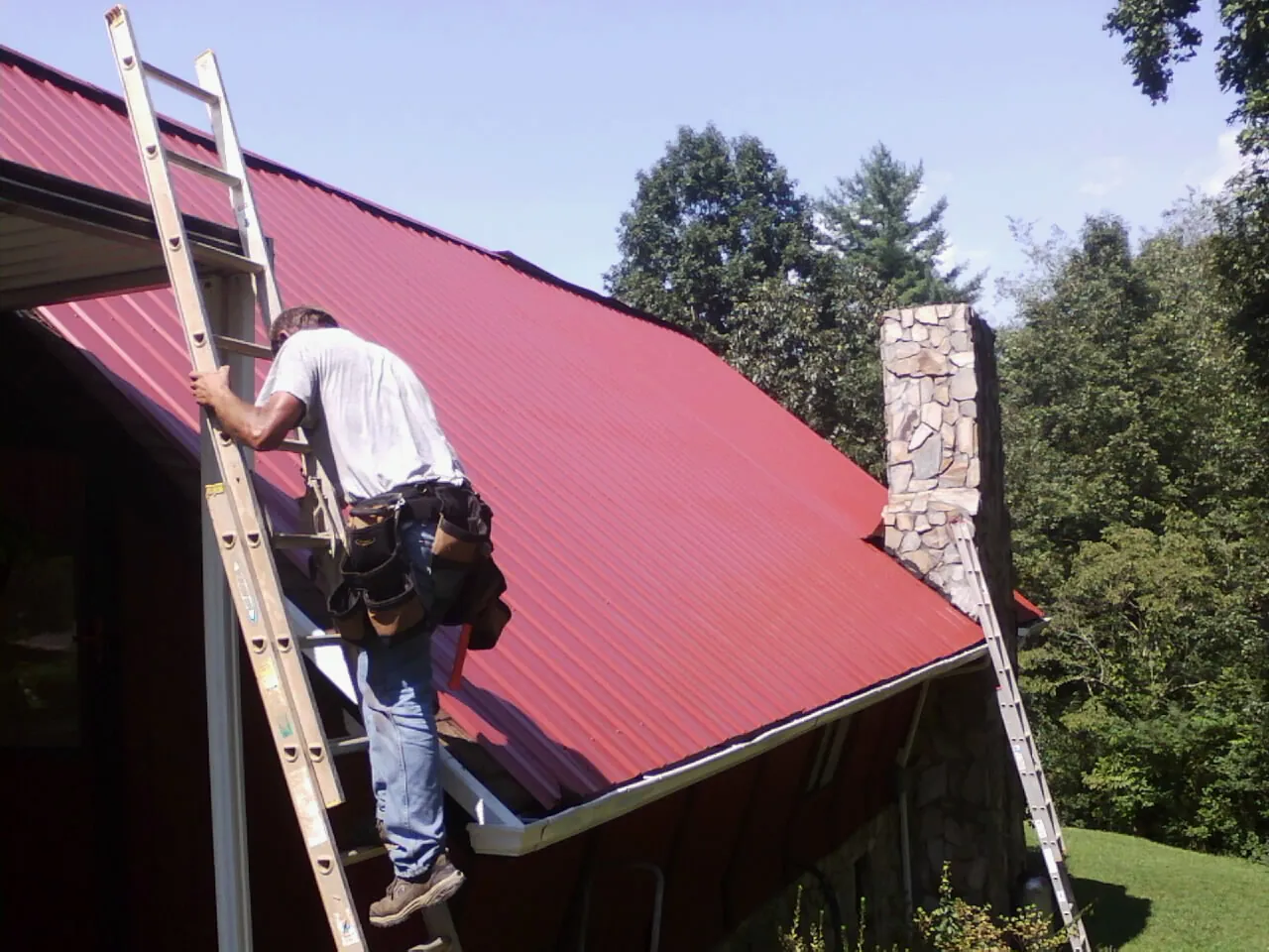 Roofer on a ladder beside a red metal roof and stone chimney — Soffit & Fascia Repair by Roofing Brothers in Mattapan